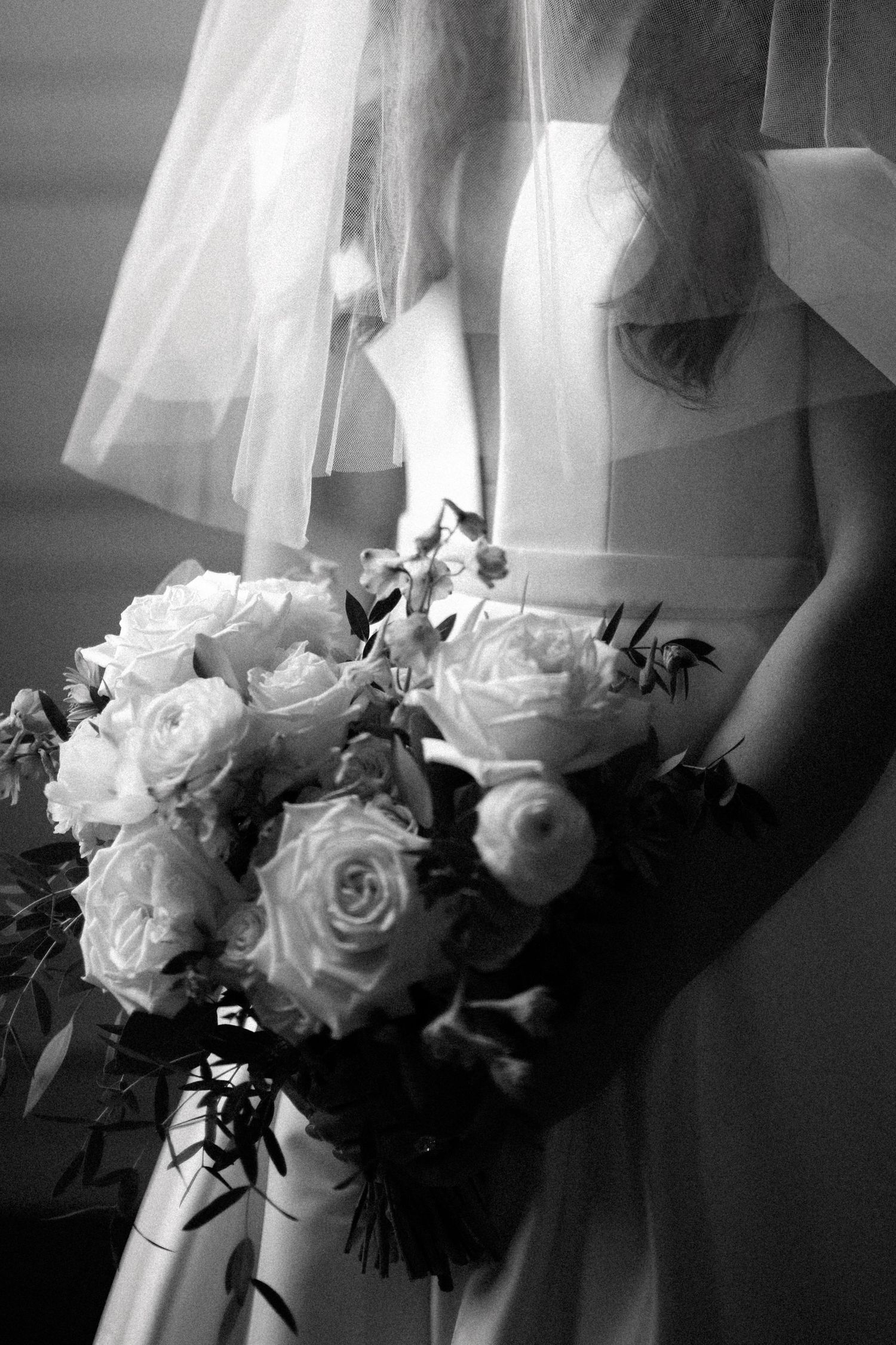 A black and white close-up of a bridal bouquet with white roses and flowing wedding dress fabric in soft focus.