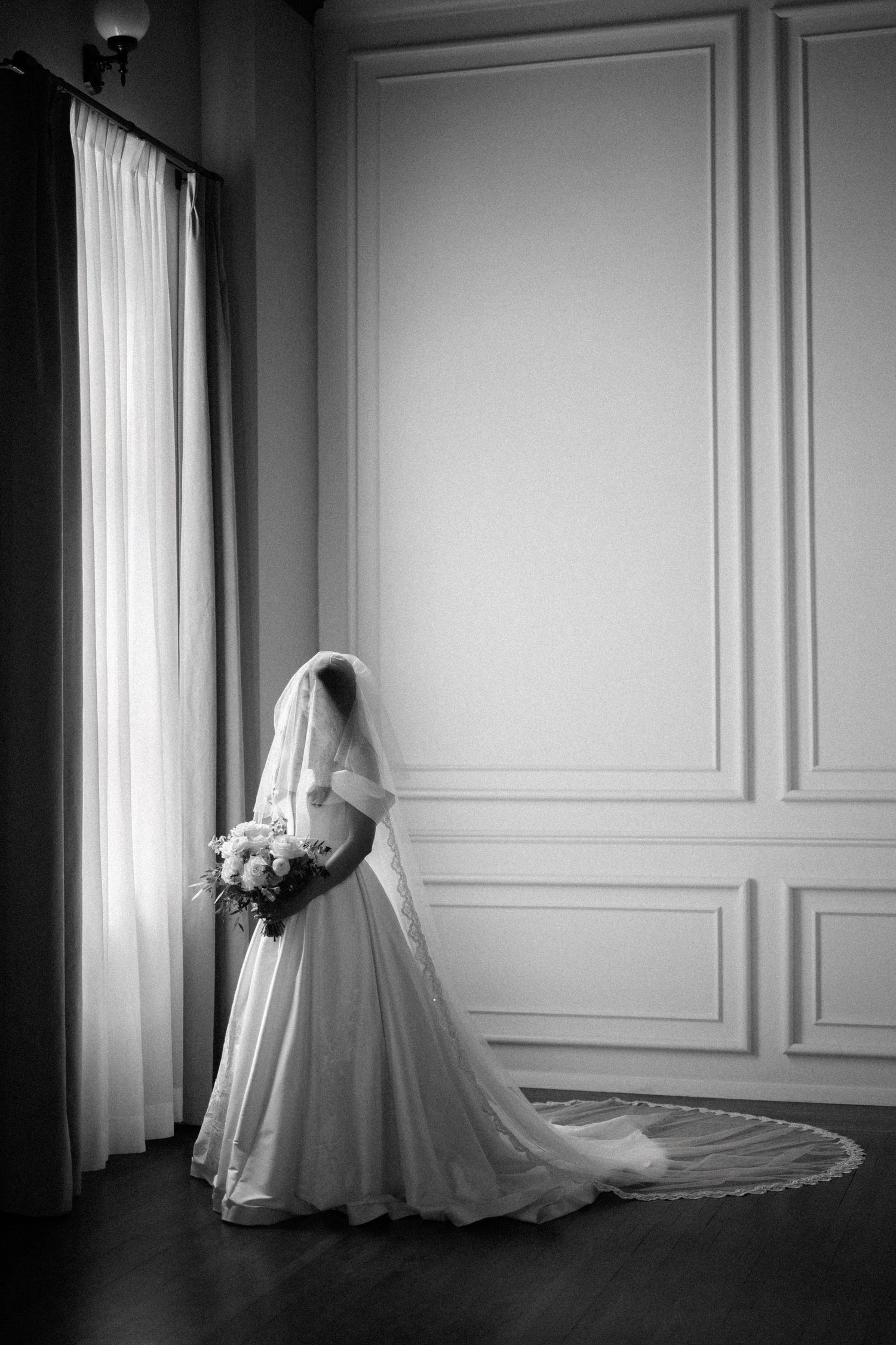 A black and white photo of an elegant wedding dress with long train and veil silhouetted against paneled walls.