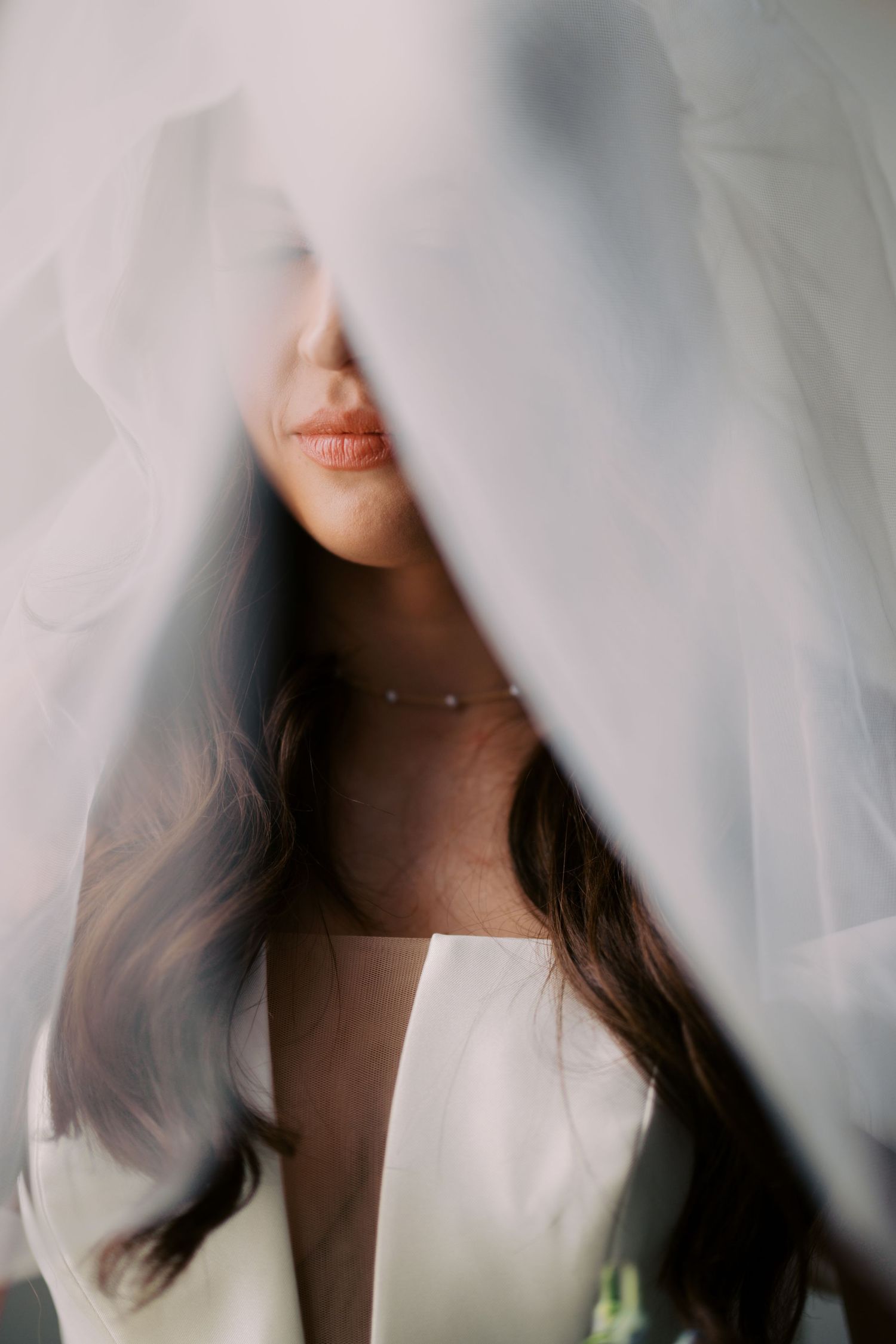 Artistic bridal portrait through sheer white veil showing soft lips and flowing dark hair.