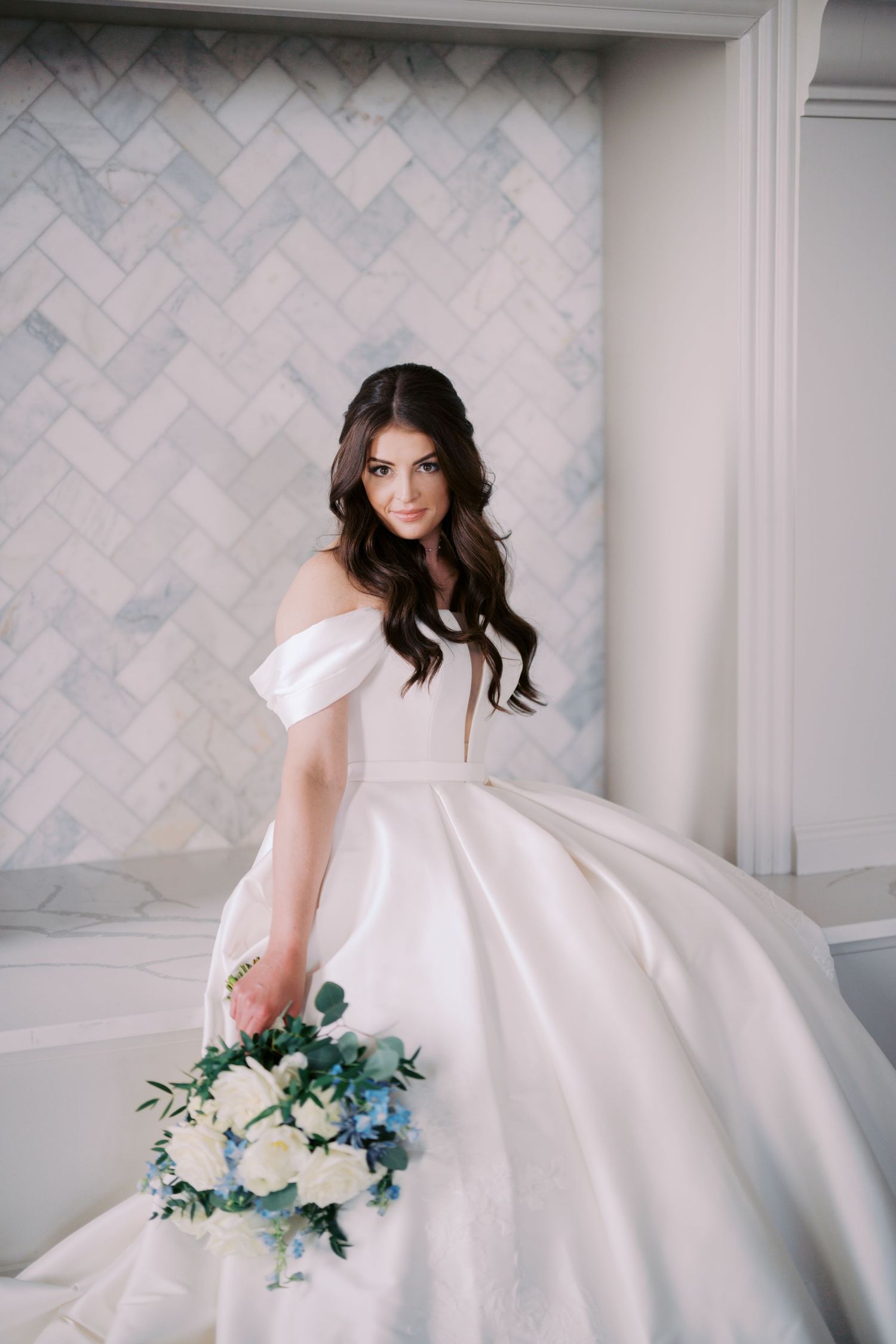 Elegant bride in off-shoulder ball gown holding blue and white bouquet against herringbone tile wall.