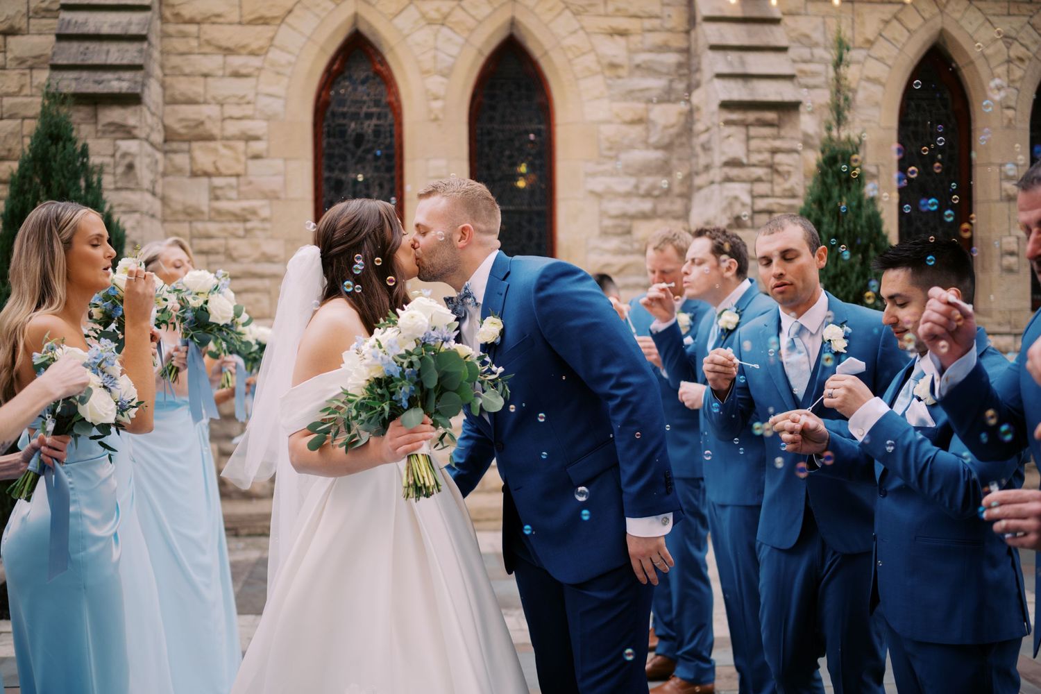 Newlywed couple shares romantic kiss while wedding party in blue dresses and suits celebrates with bubbles.