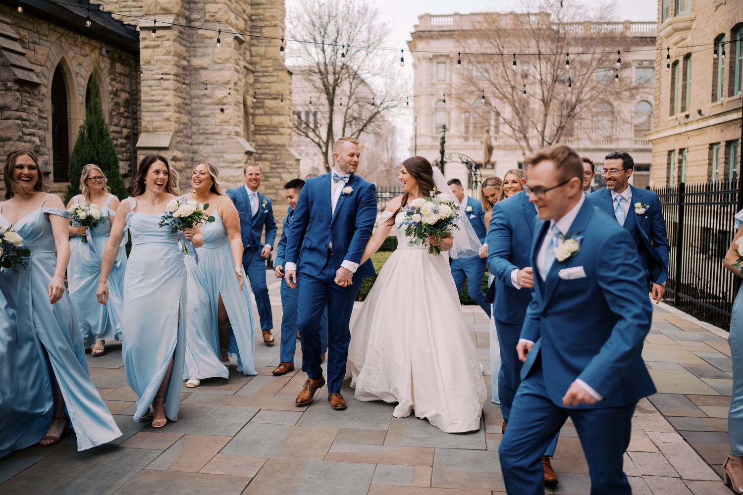 Wedding party in blue tuxedos and light blue bridesmaid dresses celebrate joyfully outside a stone church.