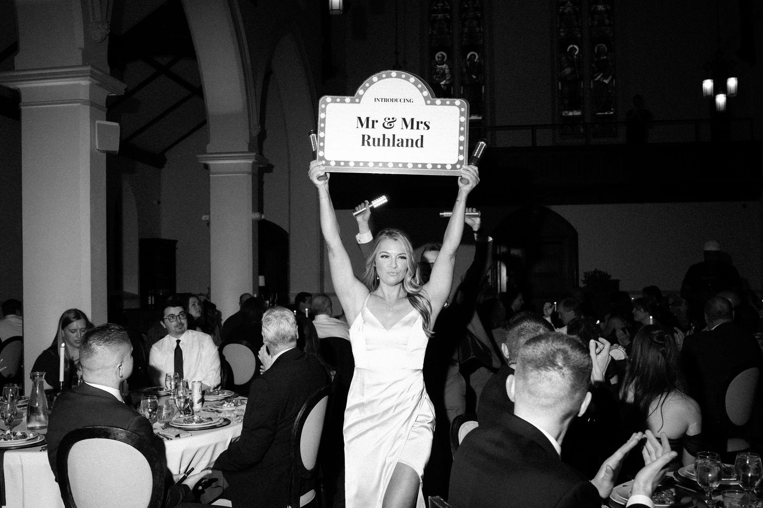 Someone in an elegant white dress holds up a sign during a formal black and white dinner event at a banquet hall.