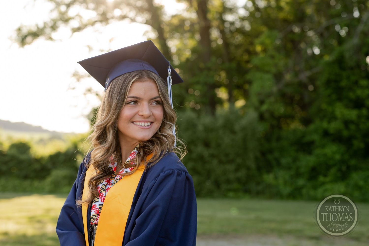 Graduate in blue and gold academic regalia poses against blurred natural background.