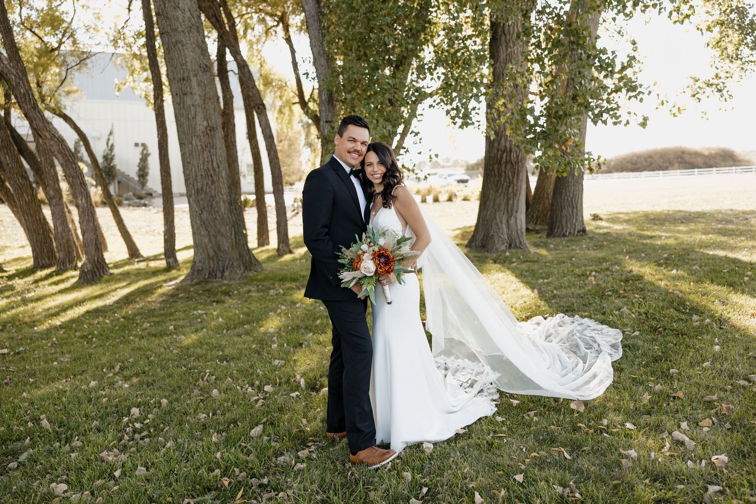 A romantic wedding portrait of newlyweds standing among trees with flowing veil in golden sunlight.