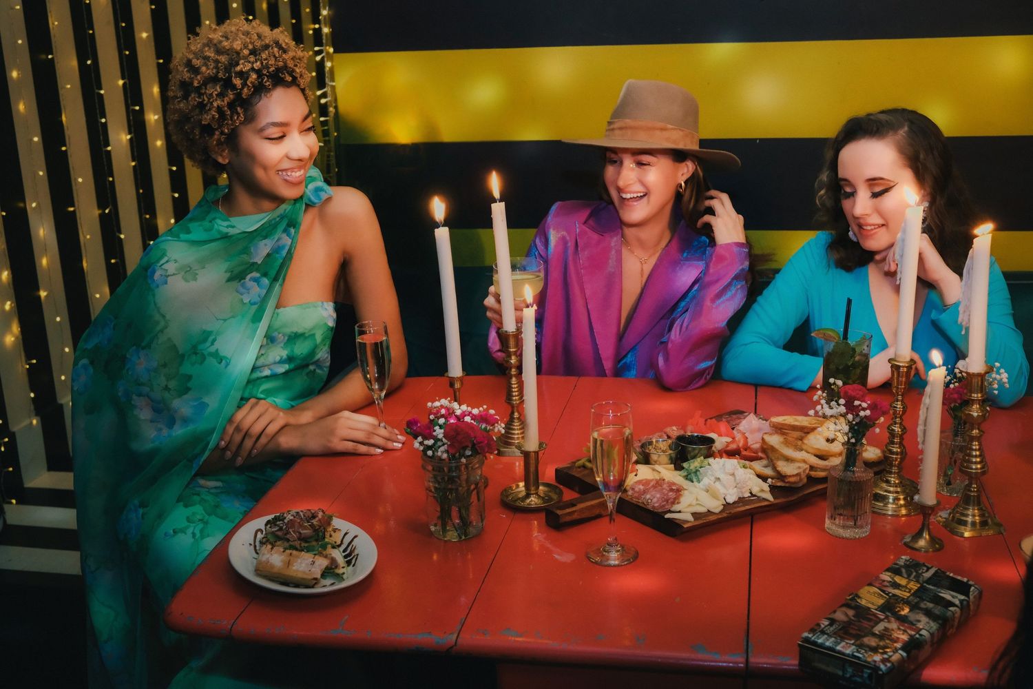 Three people in vibrant outfits sit at a red table with candlesticks and food in a moody, retro-styled restaurant setting.