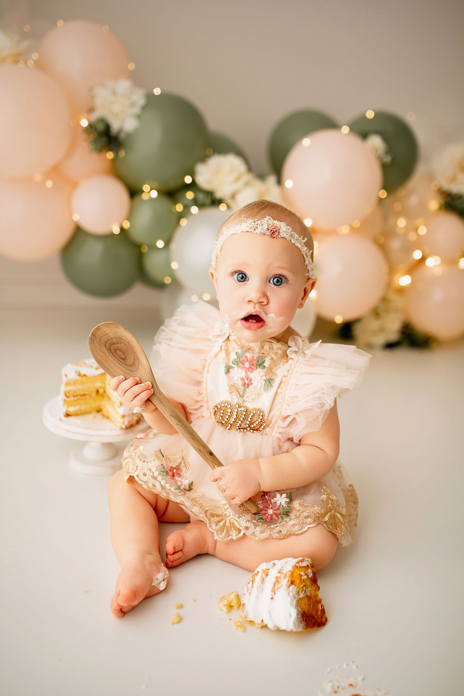 One year old baby girl with wooden spoon and sage balloon arch during cake smash session at Flowertown Photography
