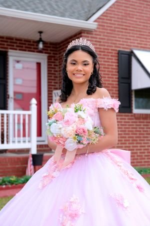 A formal portrait in front of a brick house showing a pink quinceañera gown and bouquet of pink and white flowers.