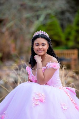 A quinceañera portrait of someone wearing a pink and white ball gown with floral details and a crystal tiara in an outdoor setting.