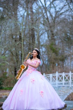Musician in flowing pink ball gown plays saxophone outdoors against wooded backdrop and white fence.