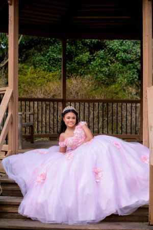 An elegant pink ball gown with floral details spreads across the wooden floor of a rustic gazebo.