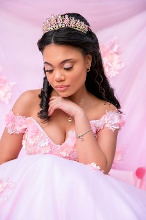 Beautiful quinceanera portrait against pink backdrop wearing sparkly crown and off-shoulder pink gown with floral details.