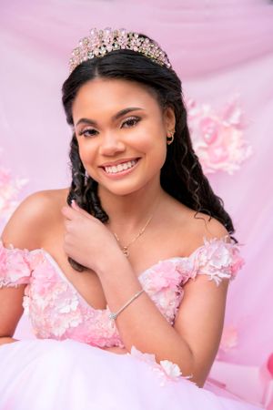 A formal portrait against a pink backdrop showing someone in an off-shoulder pink ball gown and crystal crown.