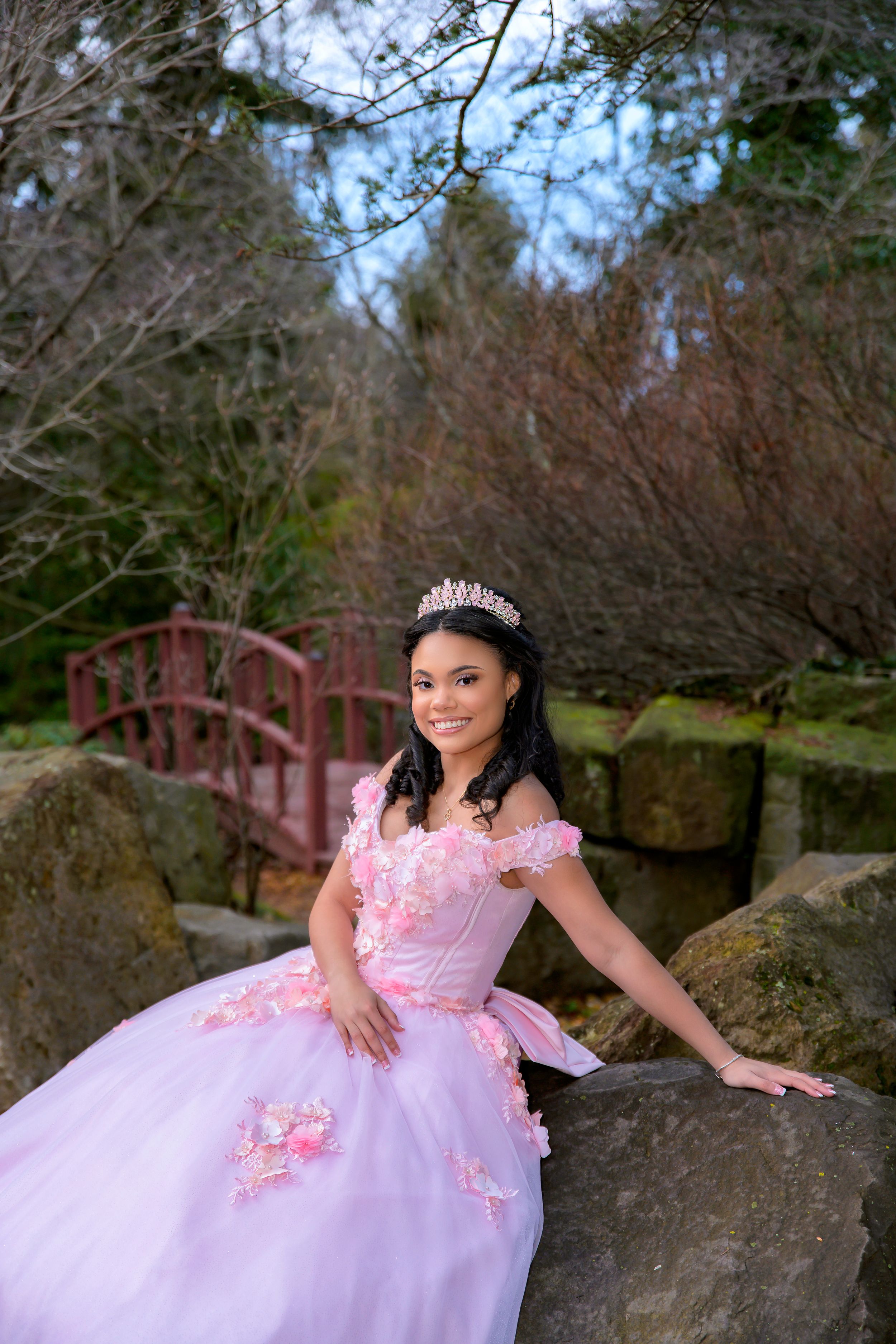 Elegant quinceanera photo in pink princess gown posing on rocks by red garden bridge in nature setting.