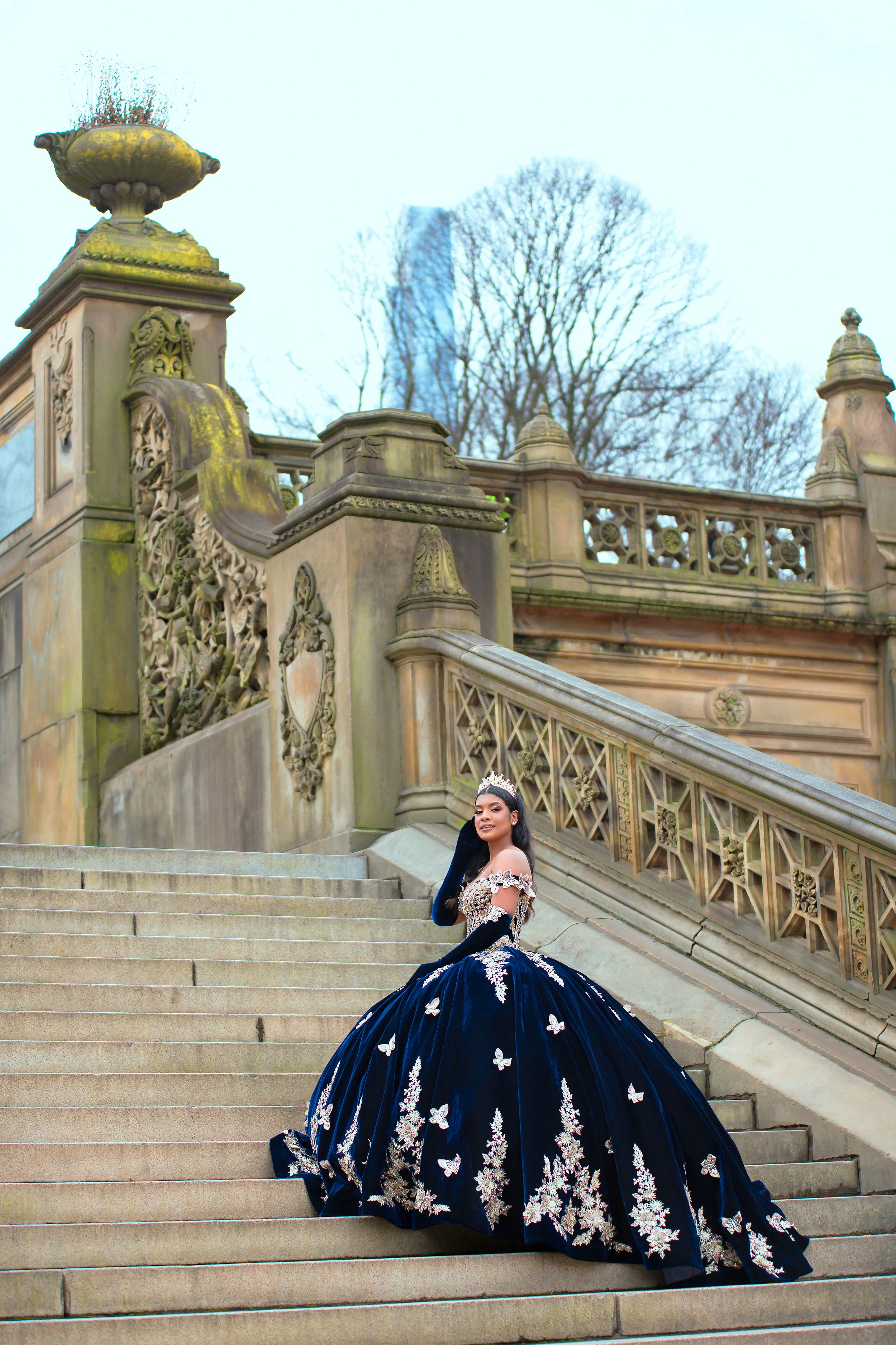 Woman in an elegant navy ball gown with floral embroidery poses on ornate stone staircase in Central Park.