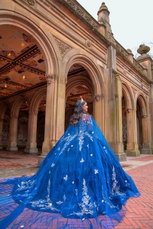 A majestic blue ballgown with butterfly embellishments flows along the arched colonnade of a historic building.