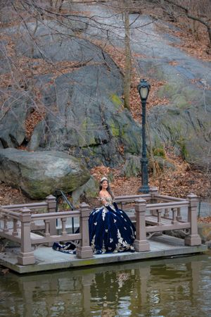 Elegant figure in a navy blue gown poses on wooden bridge deck over pond in Central Park during autumn season.