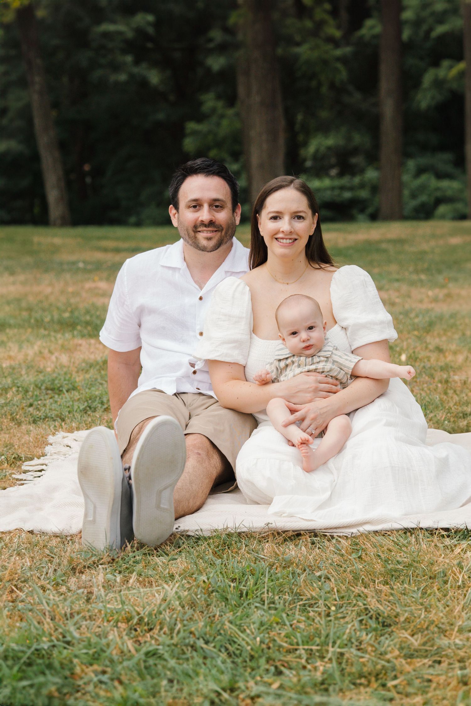 Family of three sitting cross-legged on a white blanket, smiling warmly at the camera in a green field at Barclay Farmstead.