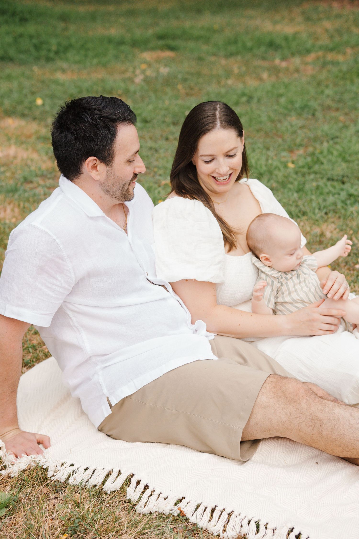 Family of three sitting on a blanket in the green field at Barclay Farmstead, sharing smiles and candid moments.