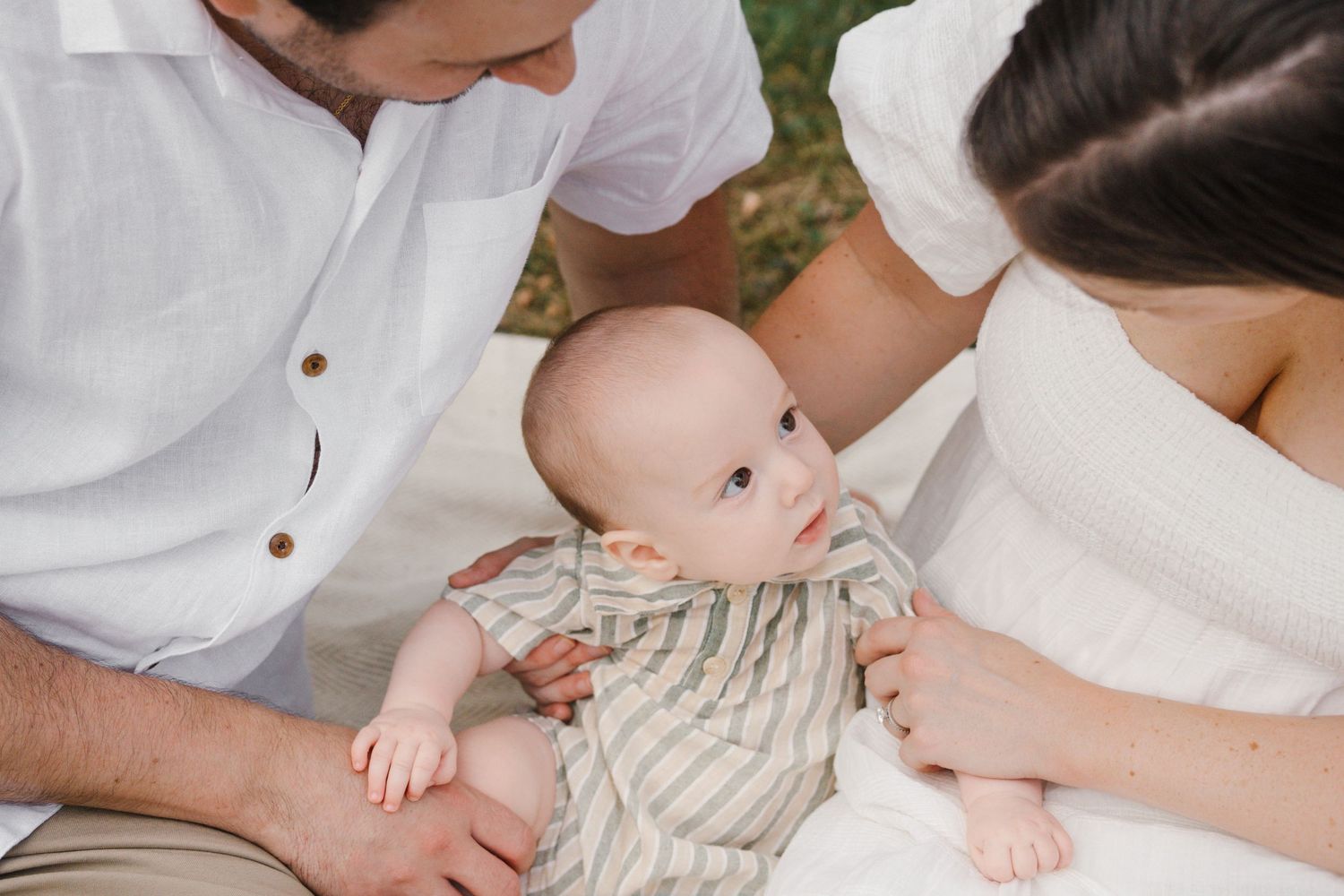 A baby sits between his parents looking up at them while posing in a green field at Barclay Farmstead.