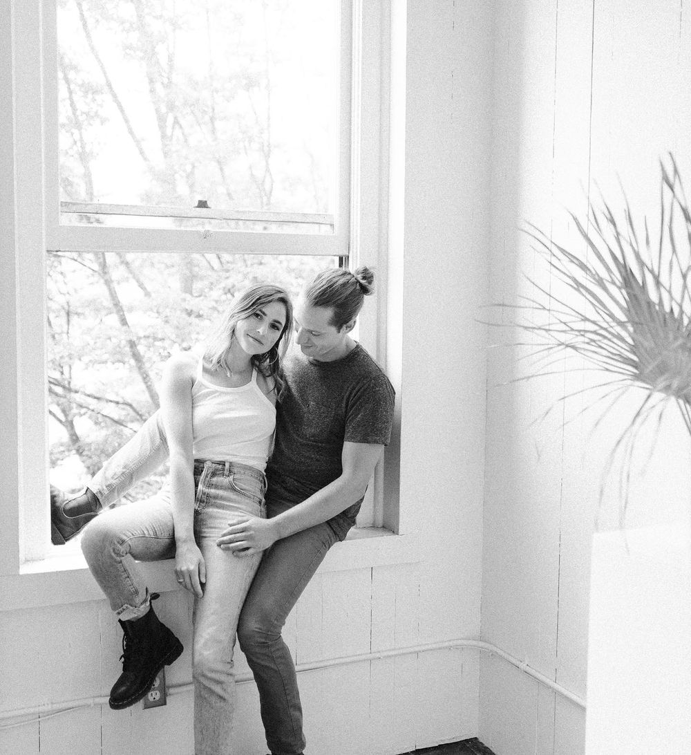 Black and white photo of a couple in casual wear leaning against a window in a minimalist bright interior.