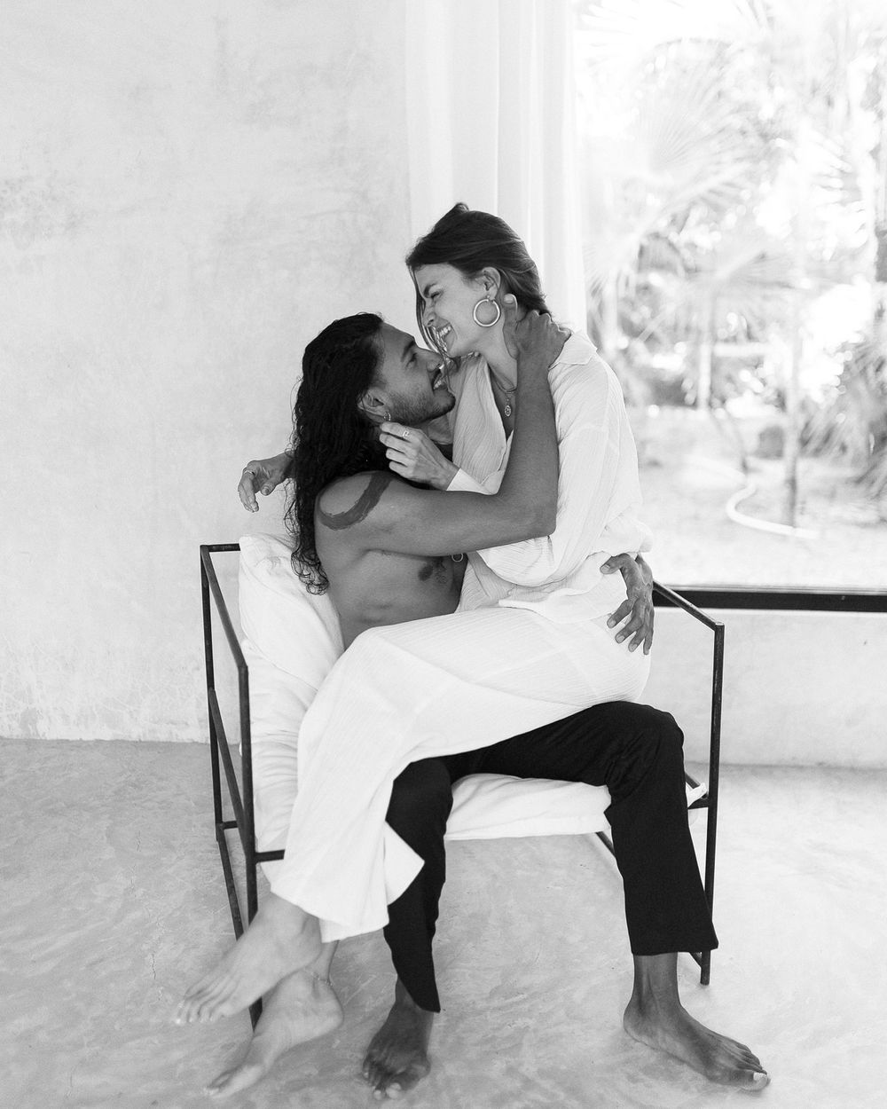 Black and white romantic photo of a couple embracing intimately while seated on a bench in a bright room.