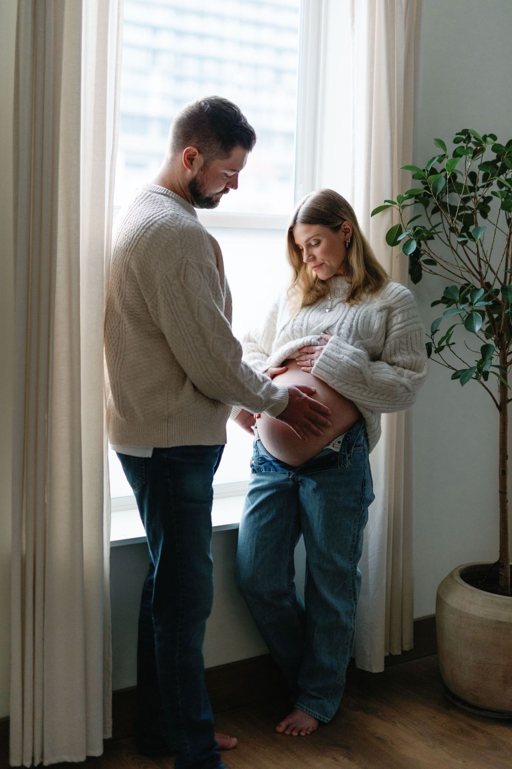 Expecting couple in cozy sweaters share tender moment by window with houseplant in natural light.