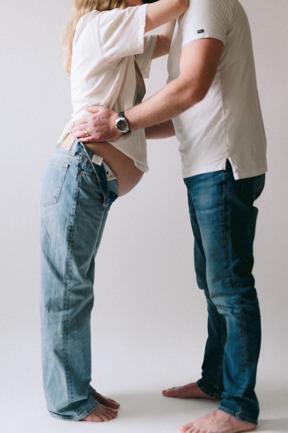 A couple in matching white tops and blue jeans stand barefoot together in an intimate embrace against a light background.