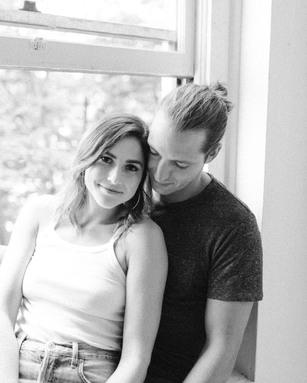 A romantic black and white photo of a couple embracing by a window with natural light streaming in.