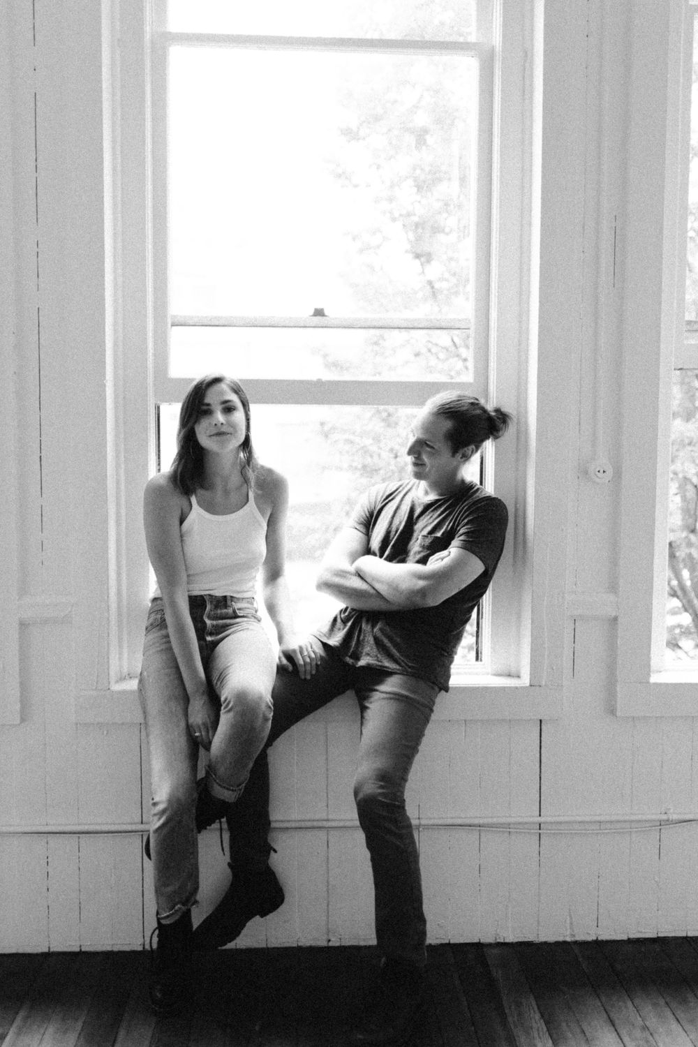 A black and white photograph of two people posing casually by a bright window in an indoor setting.