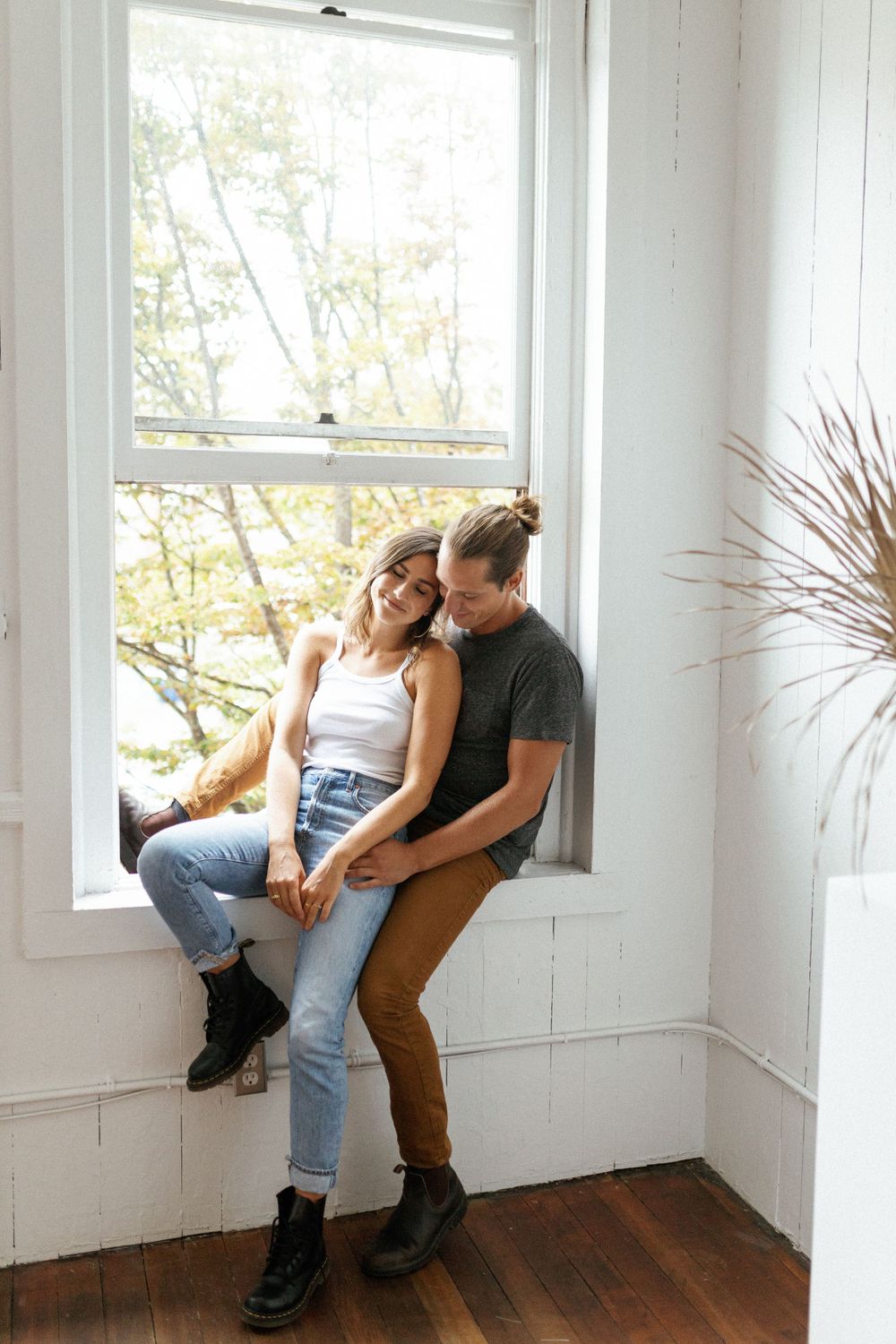 A couple sits intimately together in a bright window alcove with hardwood floors and white walls.