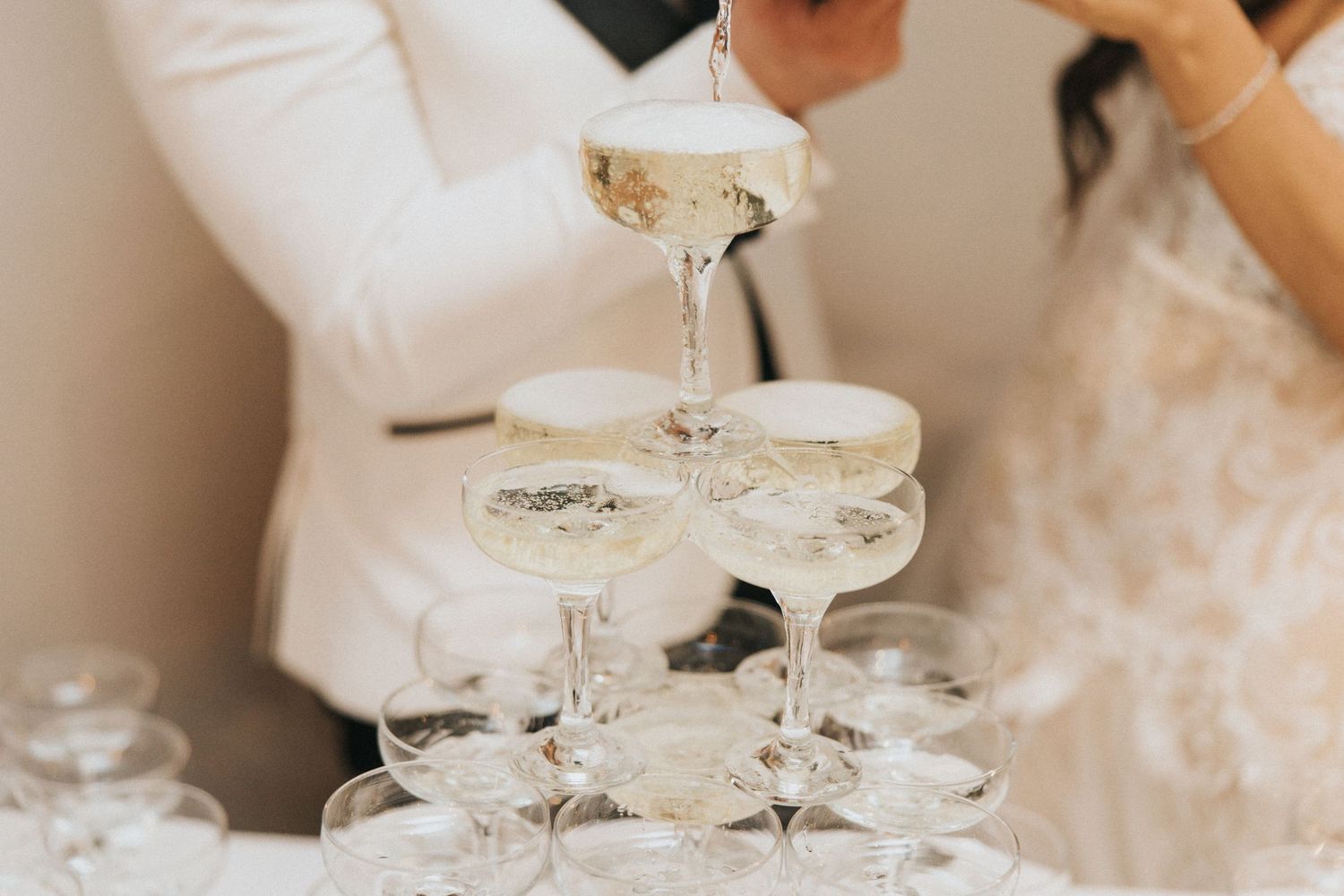 Close-up of champagne being poured into the top glass of a champagne tower, with bubbles overflowing and sparkling in the light.