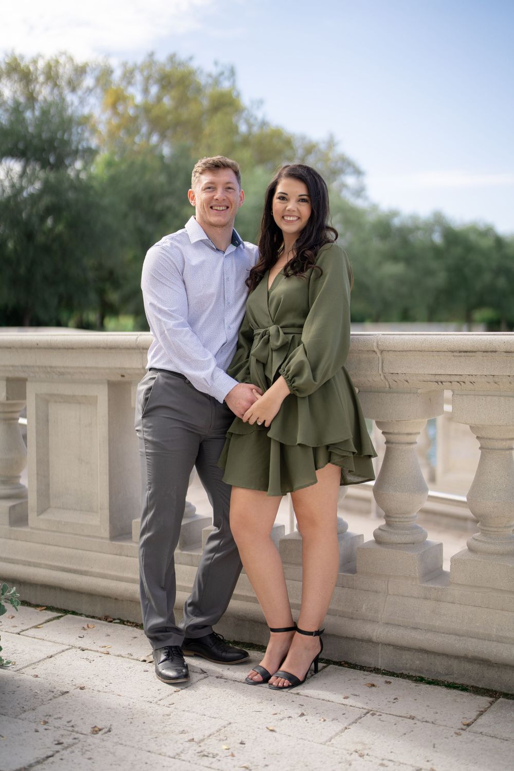 A couple poses together on a stone balcony wearing coordinated dressy outfits.
