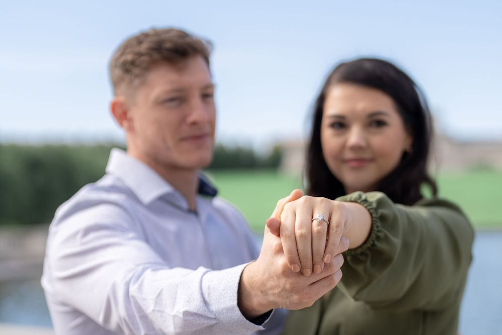 A couple shows off their engagement ring in front of a scenic water fountain backdrop.