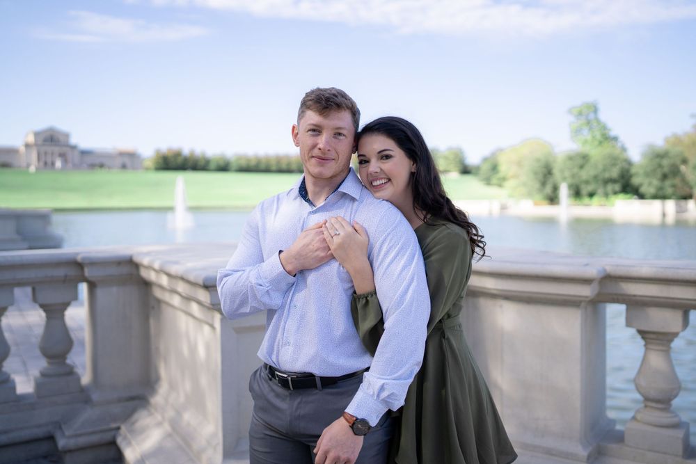A couple embraces on a stone balcony with a water fountain visible in the background.