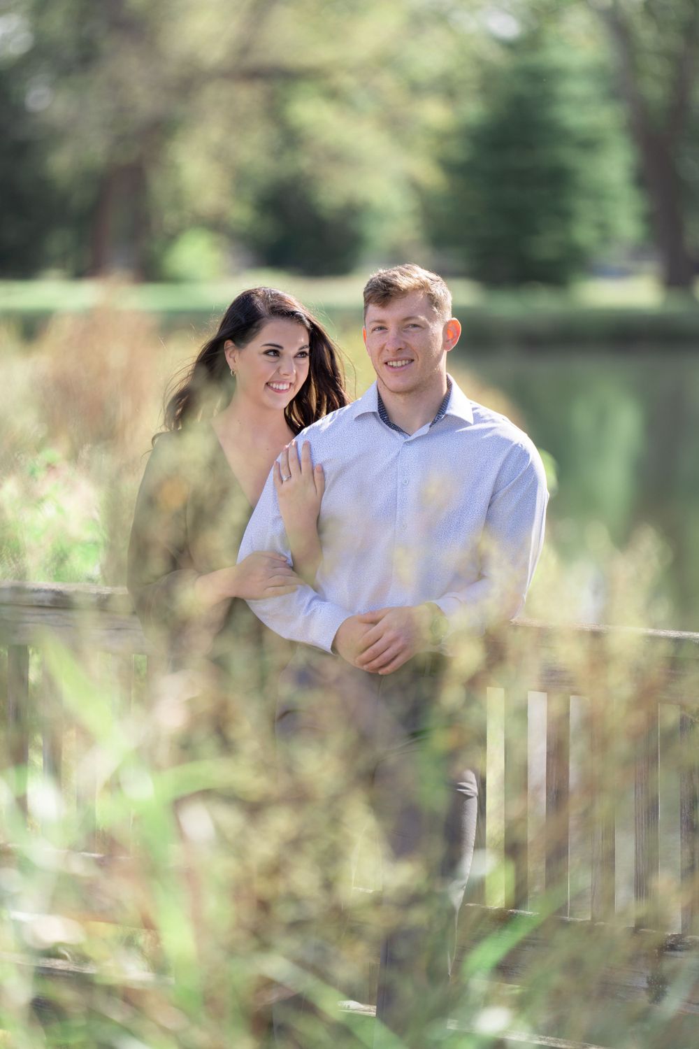 Romantic portrait through tall grass with couple standing by pond in scenic park setting.