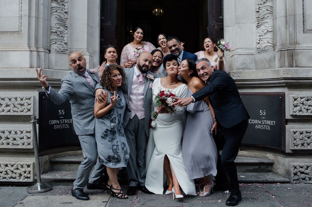 A lively wedding party poses together on stone steps outside an ornate building entrance.