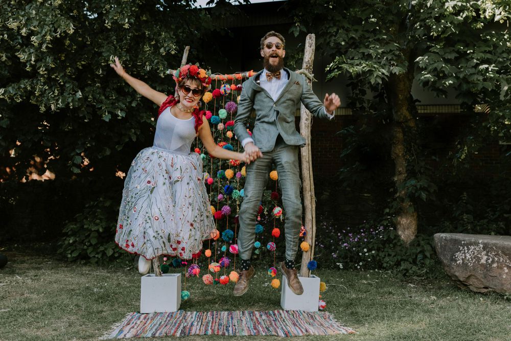 A couple stands joyfully on a floral arch in a garden setting, with the bride wearing a polka dot dress.