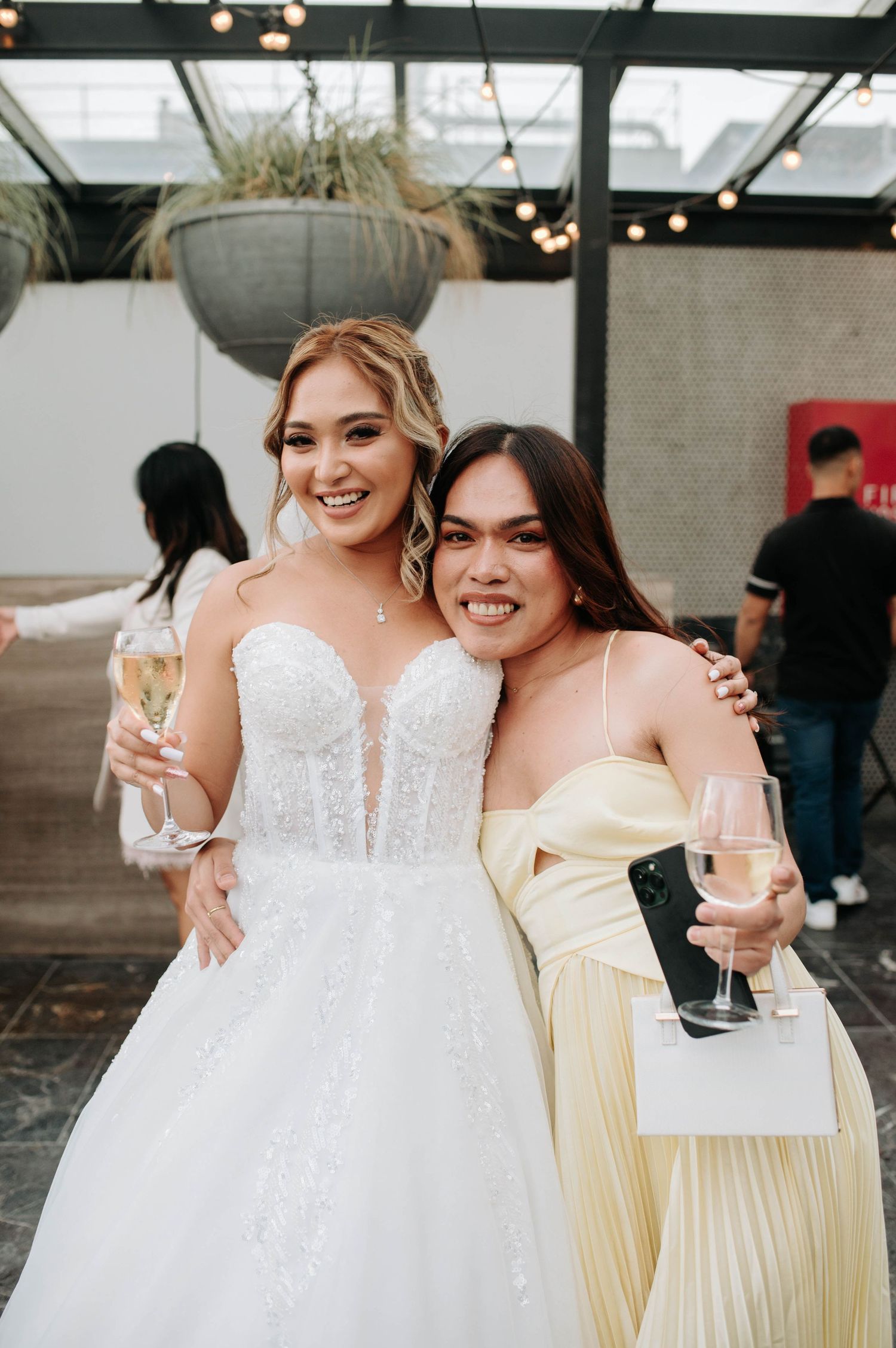 A bride in a white wedding dress stands with a guest in yellow holding champagne glasses at an indoor reception venue.