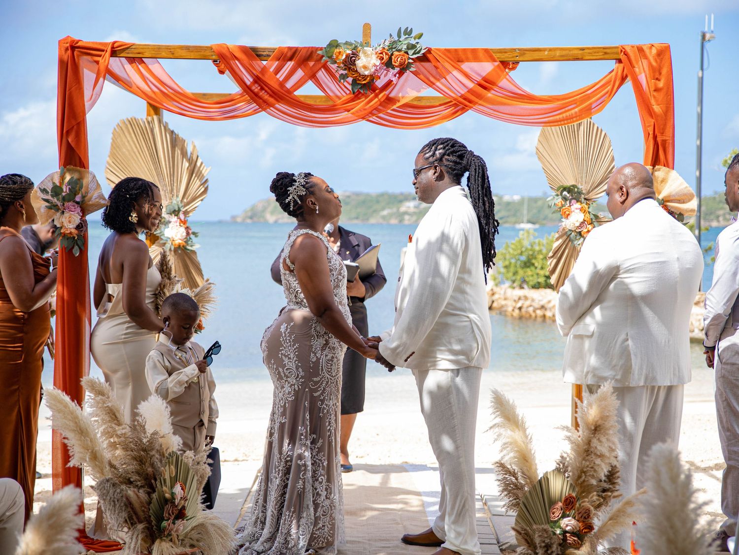 Beach wedding ceremony under orange draped arch with ocean view in background.