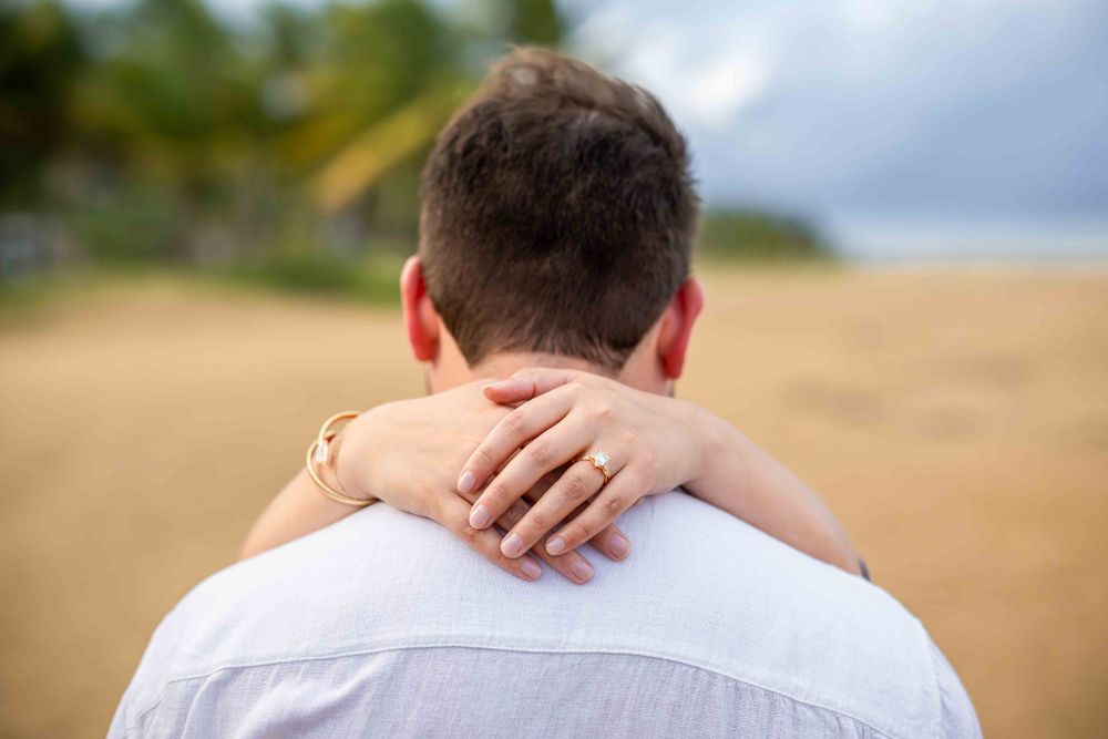 Close-up view of hands embracing from behind against a beachy background.