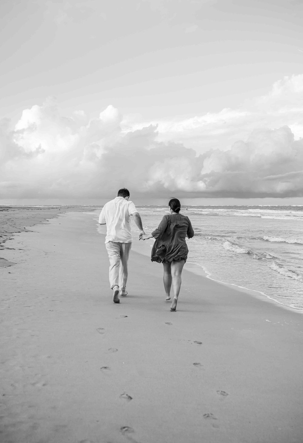 Black and white photo of a couple walking hand in hand along an empty beach.