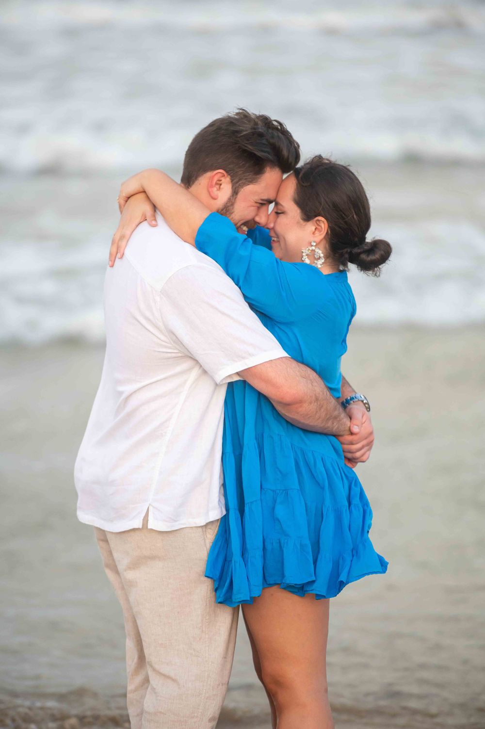 Couple in white and blue outfits embracing on a beach during a romantic photoshoot.