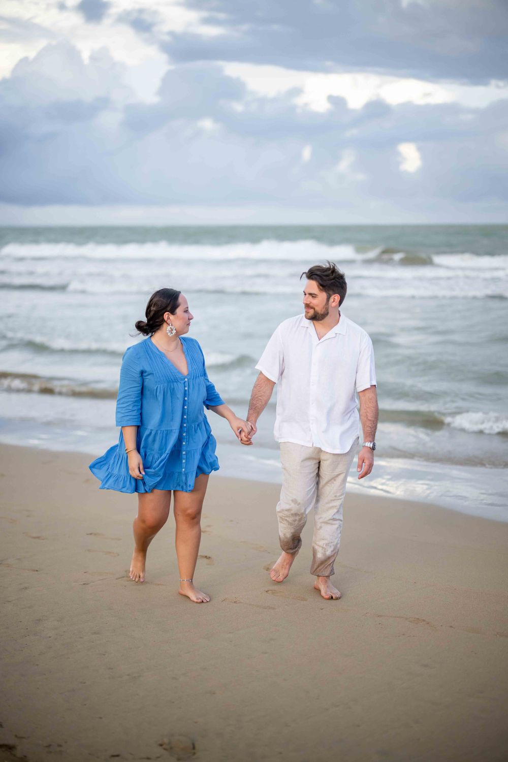 Playful couple walking and laughing together along the shoreline on an overcast beach day.