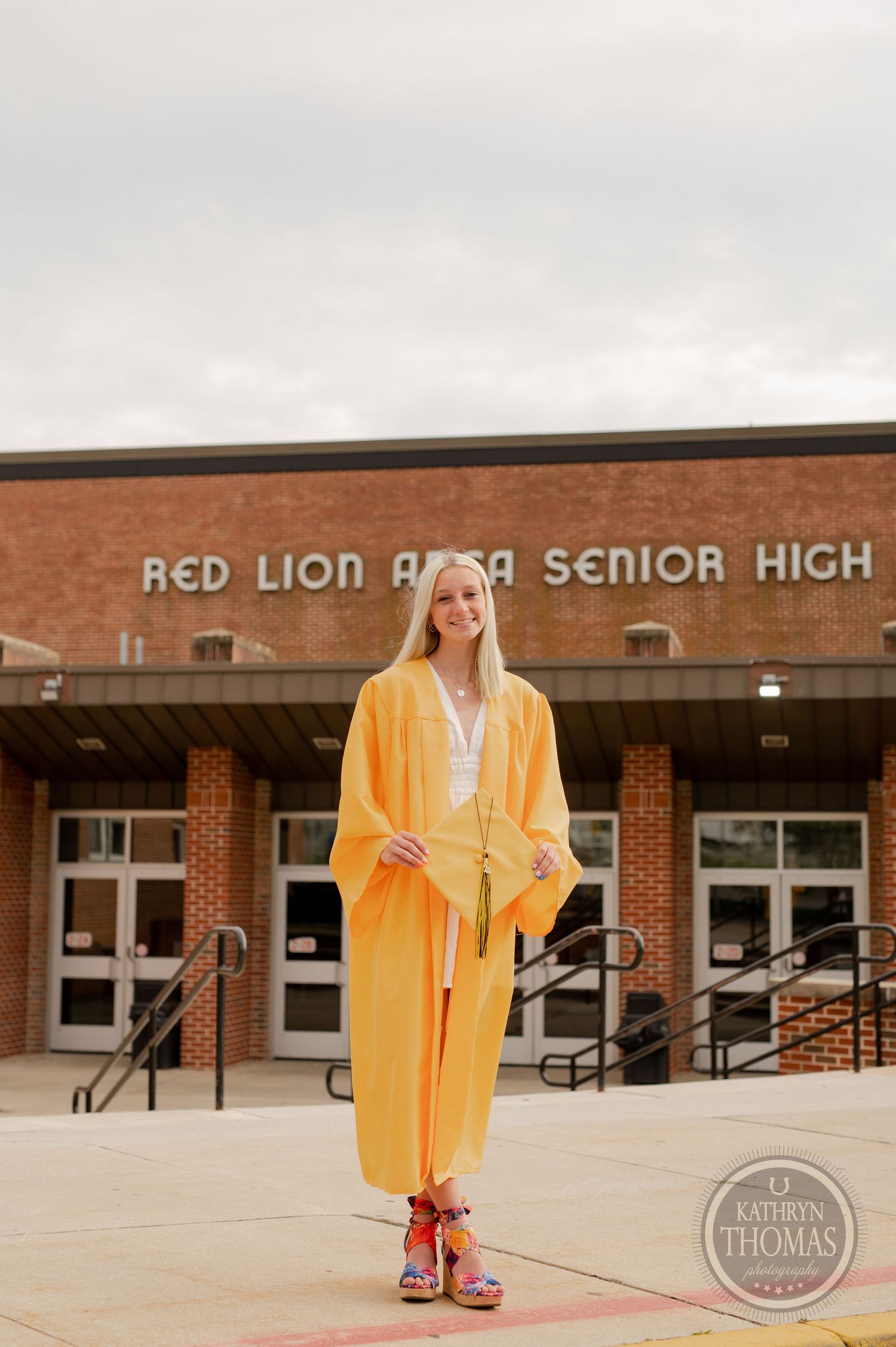 Graduate wearing yellow graduation attire and colorful shoes stands in front of brick school building.