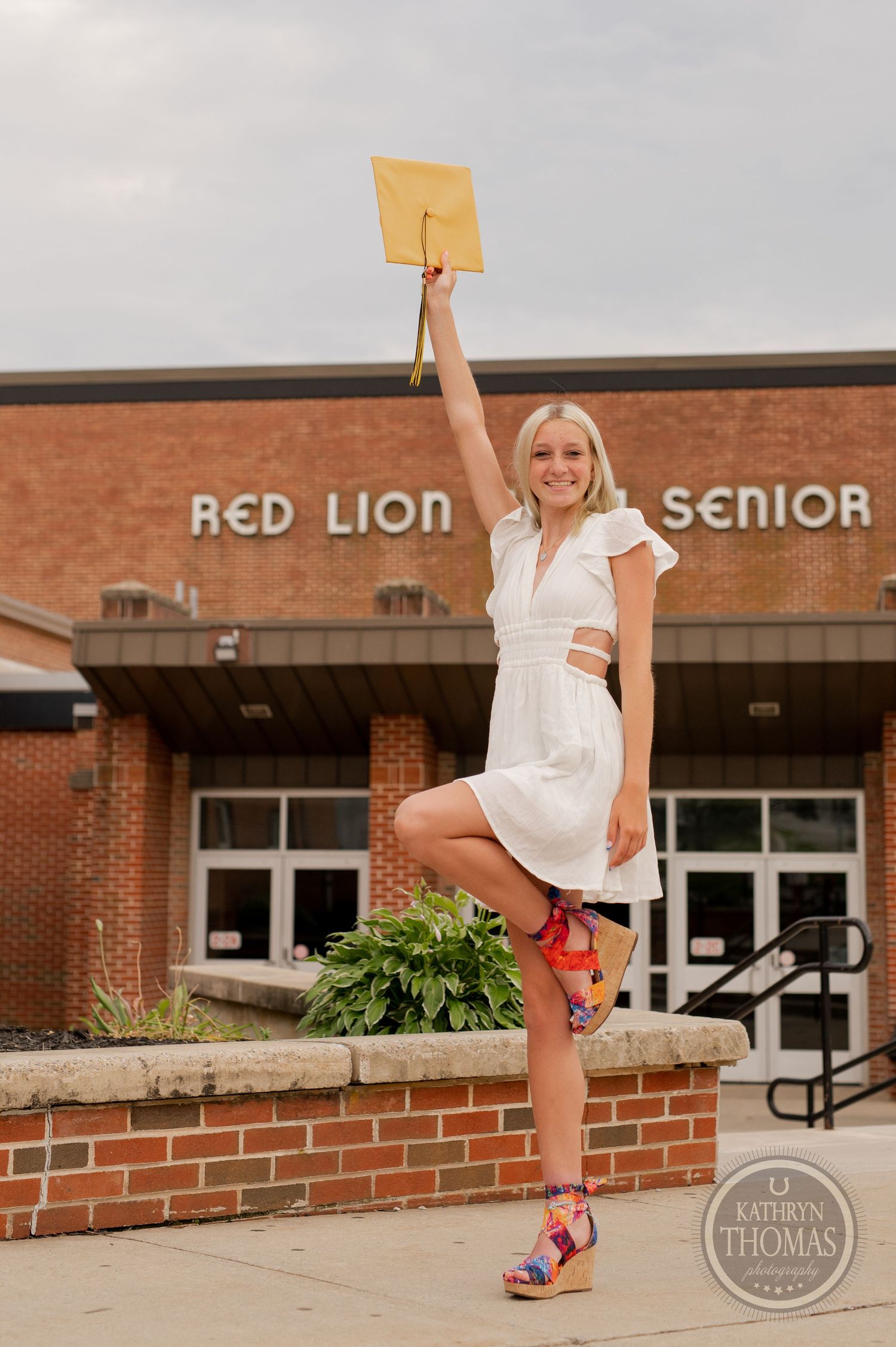 Graduate in white dress holds diploma high while posing with one leg raised outside school.