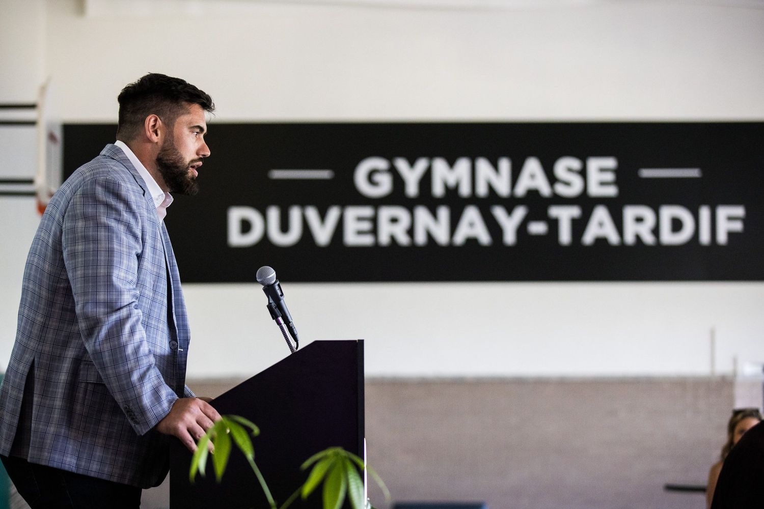 Homme chic bien connu donnant un discours dans le gymnase d'une école secondaire avec en arrière-plan une affiche portant son nom.