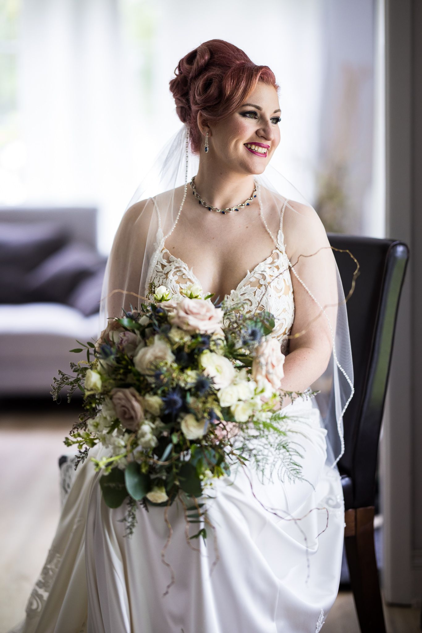 Une mariée souriante en robe blanche pose assise avec un élégant bouquet de fleurs. Une lumière douce et naturelle l'éclaire.