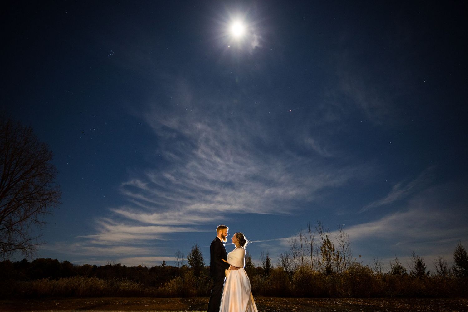 Un couple romantique s'enlace sous une pleine lune éclatante, avec de légers nuages ​​dans le ciel nocturne. Un flash de caméra les éclaire.