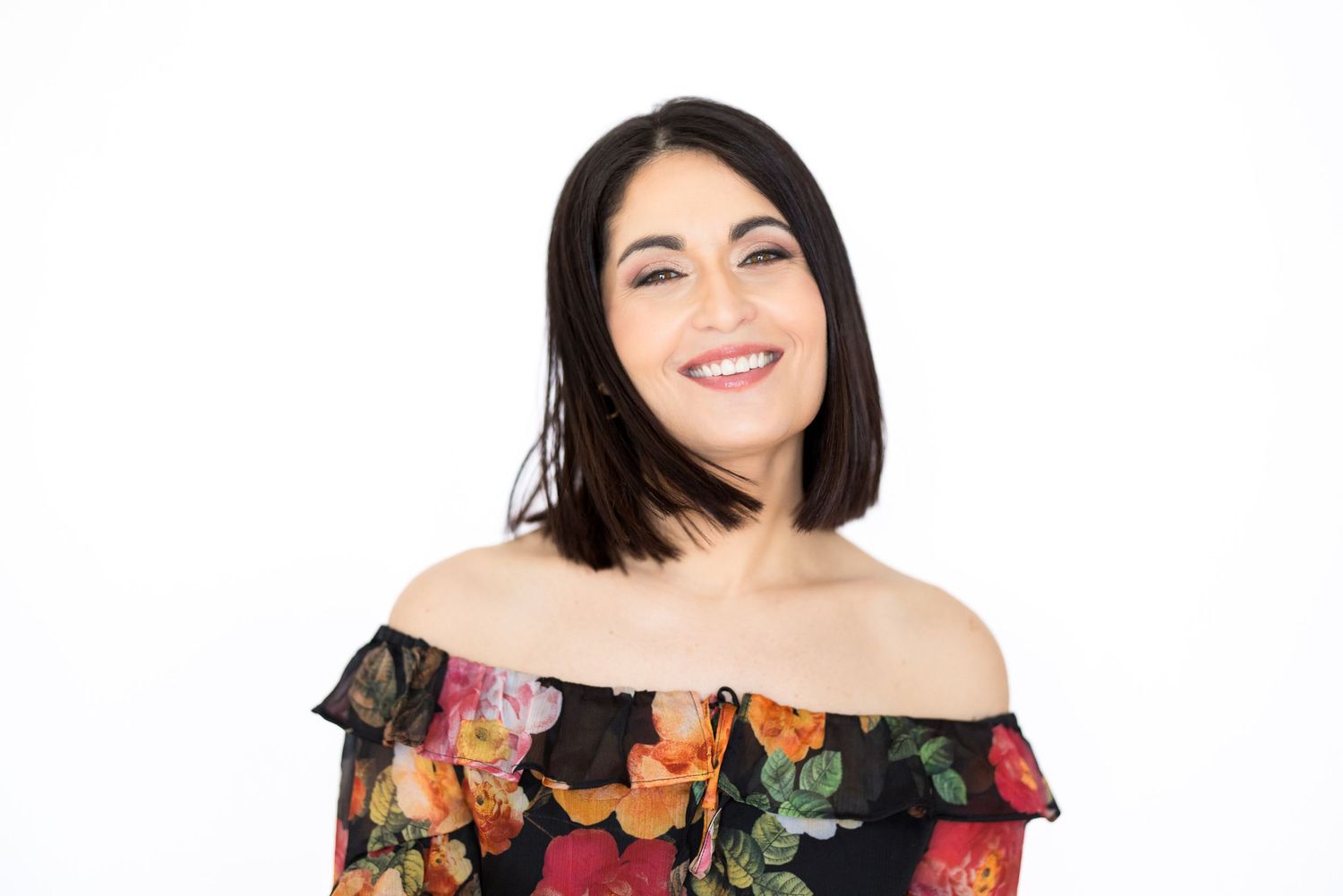 Portrait professionnel d'une femme en robe fleurie à épaules dénudées qui pose devant un fond blanc avec un sourire éclatant.